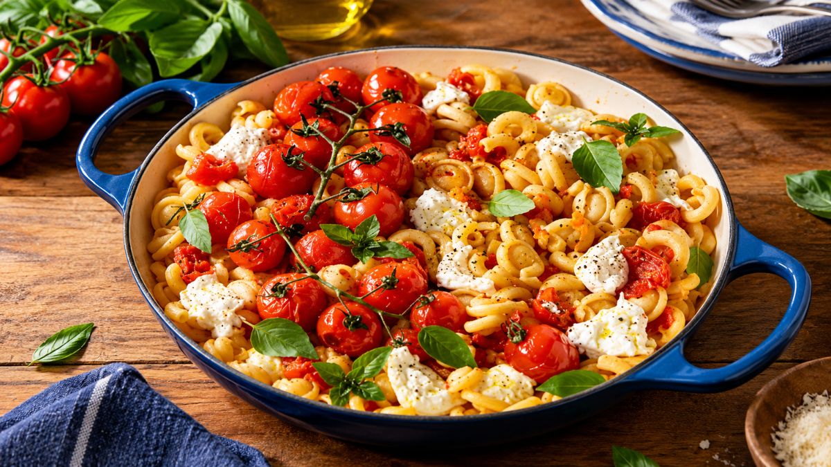 Uma panela de cerâmica azul cheia de massa artesanal picante com queijo burrata cremoso e tomates cherry no ramo assados, guarnecida com folhas de manjericão fresco.