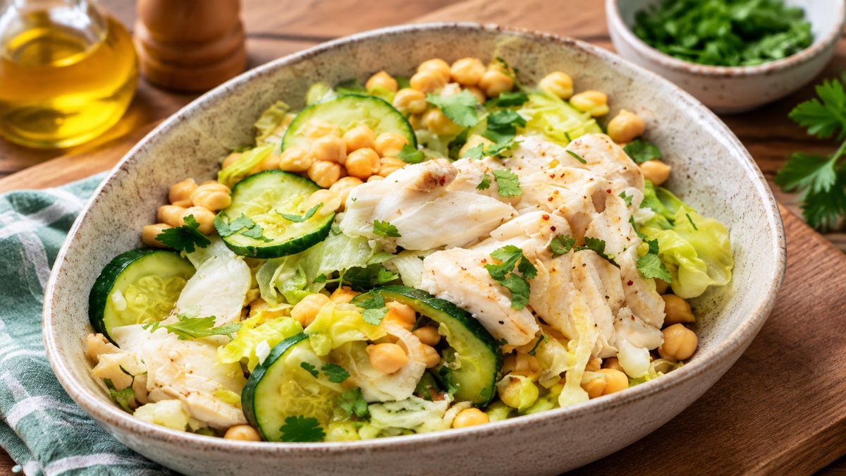 Uma tigela rústica cheia de salada de bacalhau cozido com grão-de-bico, fatias de curgete e couve fresca, polvilhada com salsa e coentros picados.