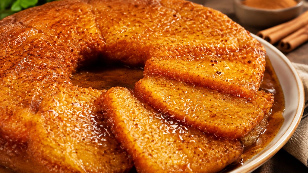 Um plano próximo de uma travessa de cerâmica contendo a tradicional sopa dourada conventual portuguesa. Fatias de pão de ló embebidas numa calda de açúcar dourada e brilhante estão cobertas com um doce cremoso de gemas, amêndoas e chila, polvilhadas com canela. Algumas fatias estão cortadas e sobrepostas. No fundo, vêem-se paus de canela desfocados sobre uma mesa de madeira.