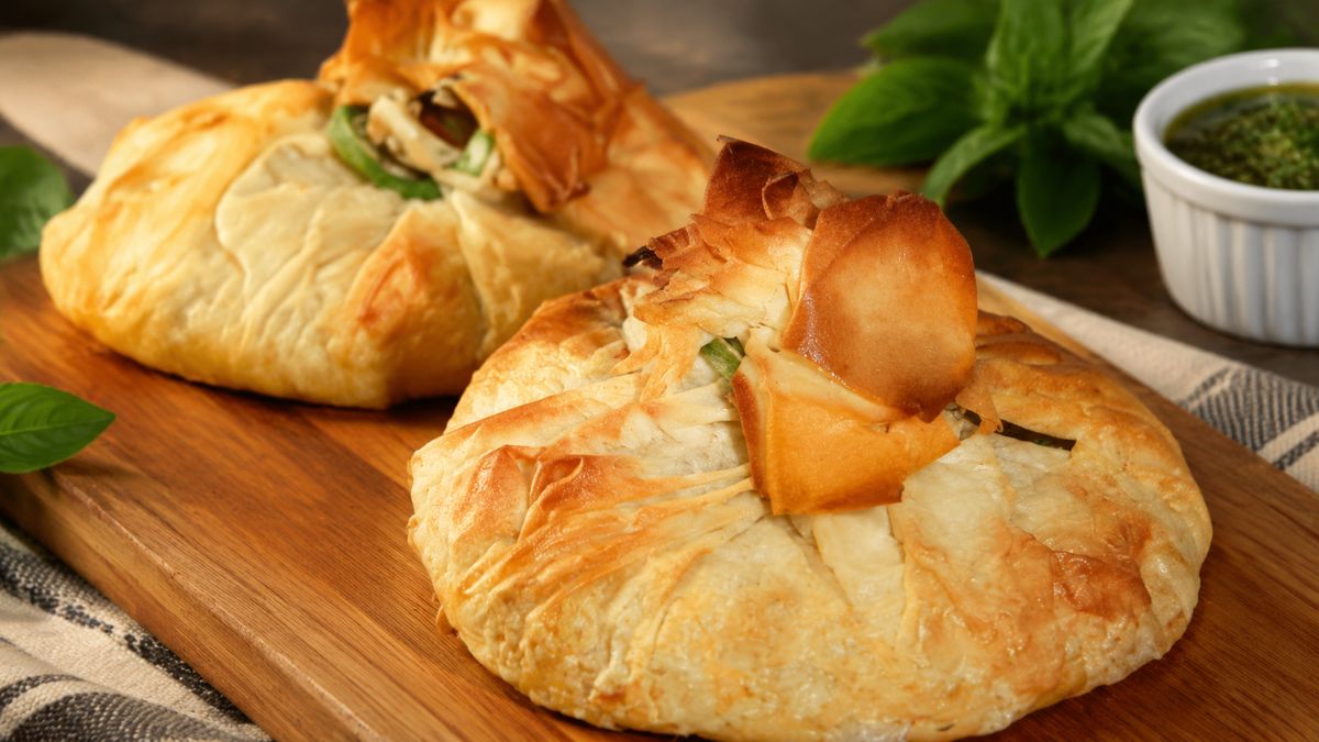 Duas trouxas de bacalhau douradas e crocantes, feitas com massa filo ou folhada, servidas numa tábua de madeira com molho pesto e manjericão fresco ao fundo.