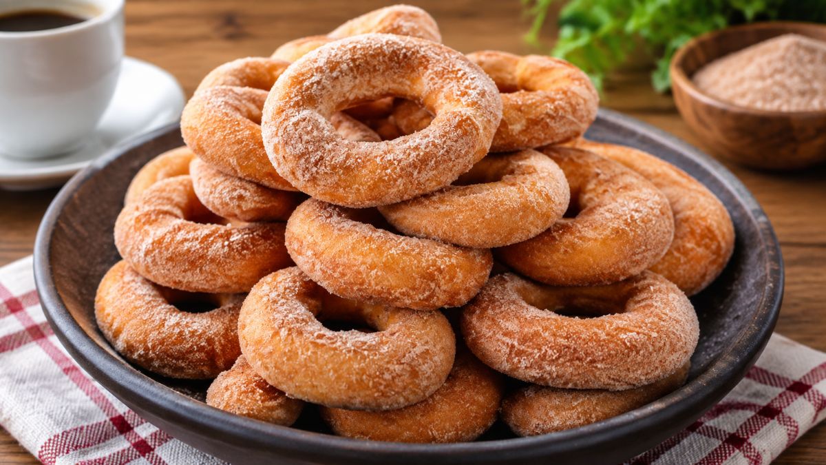 rosquinhas fritas redondas cobertas com açúcar e canela empilhadas em uma tigela de cerâmica preta rústica, com uma xícara de café ao fundo