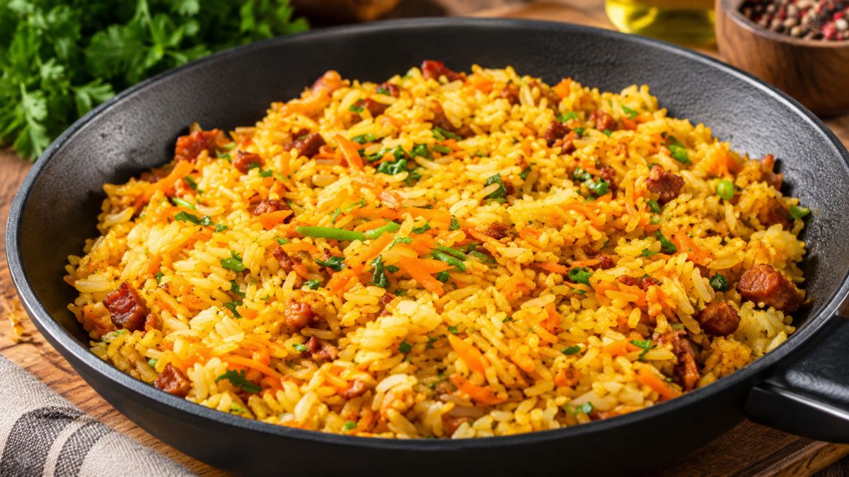 Fotografia de um apetitoso arroz de cenoura soltinho preparado numa frigideira preta com pequenos pedaços de chouriço e salsa fresca salpicada por cima.