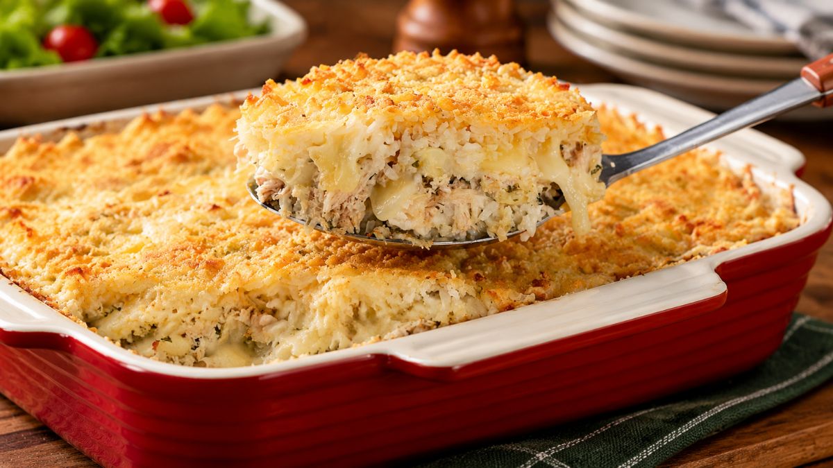 Uma travessa vermelha cheia de arroz de forno cremoso com frango desfiado gratinado com queijo derretido e uma colher de servir tirando uma porção.