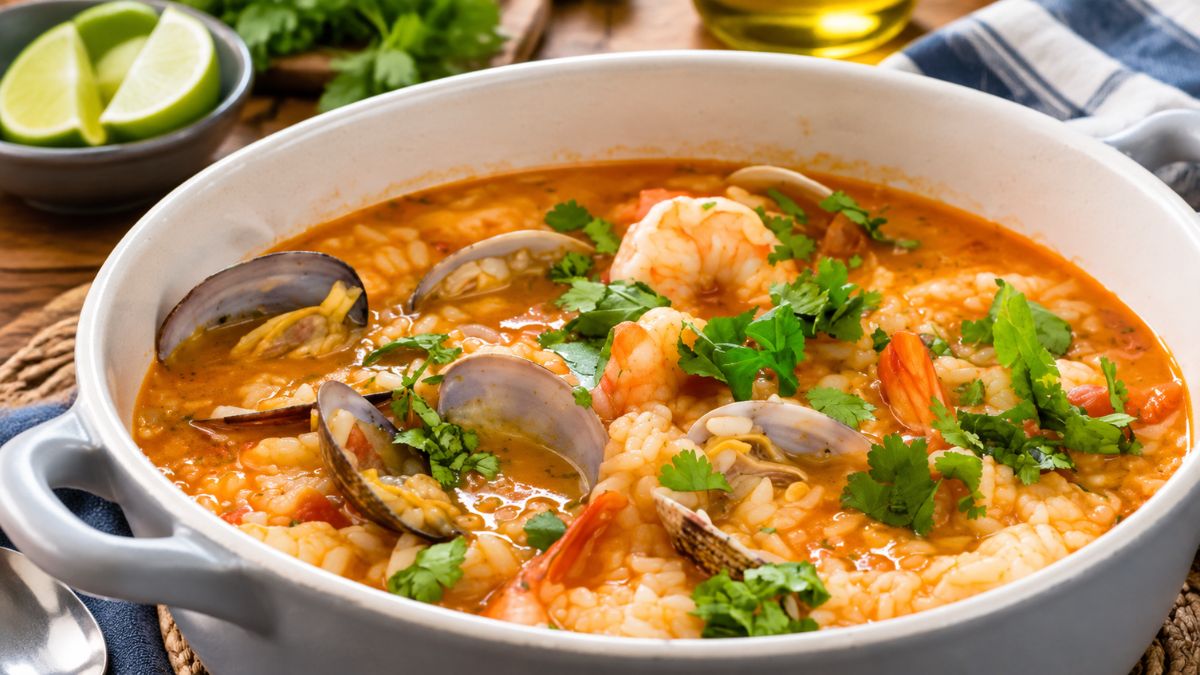 Uma panela rústica cinzenta cheia de arroz malandrinho de tamboril, decorado com amêijoas abertas na concha, camarões suculentos e coentros frescos picados. Ao fundo, gomos de lima e azeite num ambiente de cozinha acolhedor.