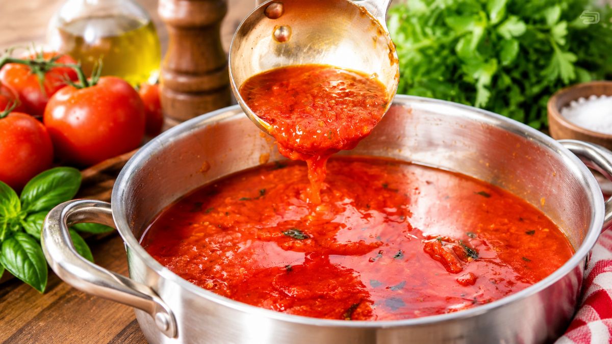 Concha de metal servindo molho de tomate caseiro fresco em uma panela com tomates, manjericão e azeite ao fundo.
