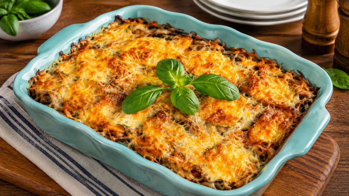 Uma travessa de cerâmica azul-turquesa preenchida com um gratinado de abobrinha e queijo derretido dourado, decorada com manjericão fresco, sobre uma tábua de madeira.