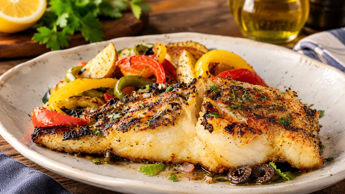 Lombo de bacalhau grelhado com lascas douradas, servido com legumes assados coloridos, pimentos, batatas e azeitonas pretas.