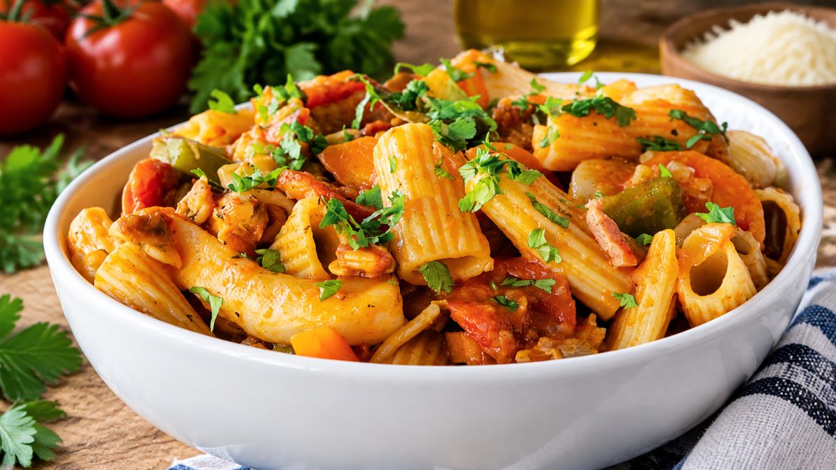 Prato fundo branco servido com massada de pota e berbigão macio, macarrão riscado envolto em molho de tomate aromático com pimentos e coentros frescos polvilhados.