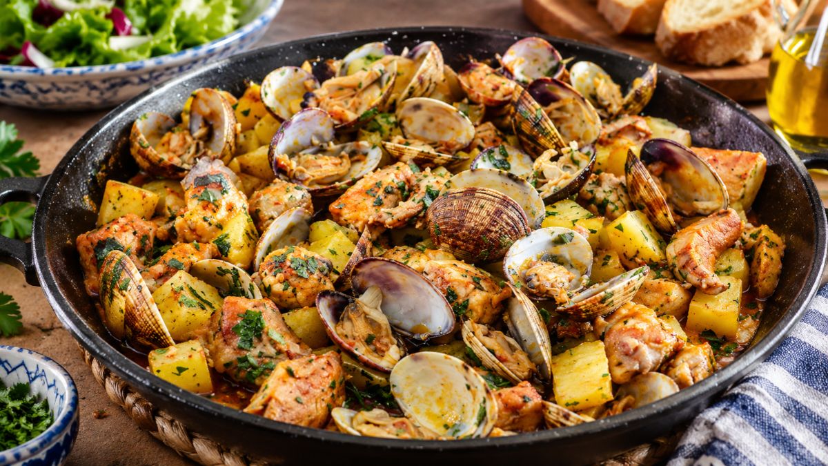 Vista aproximada de Carne de Porco à Alentejana tradicional servida numa frigideira de ferro fundido preta. O prato contém cubos de carne de porco suculento, batatas fritas crocantes e amêijoas abertas polvilhadas com coentros frescos picados. Ao fundo, vê-se uma salada fresca e fatias de pão rústico.