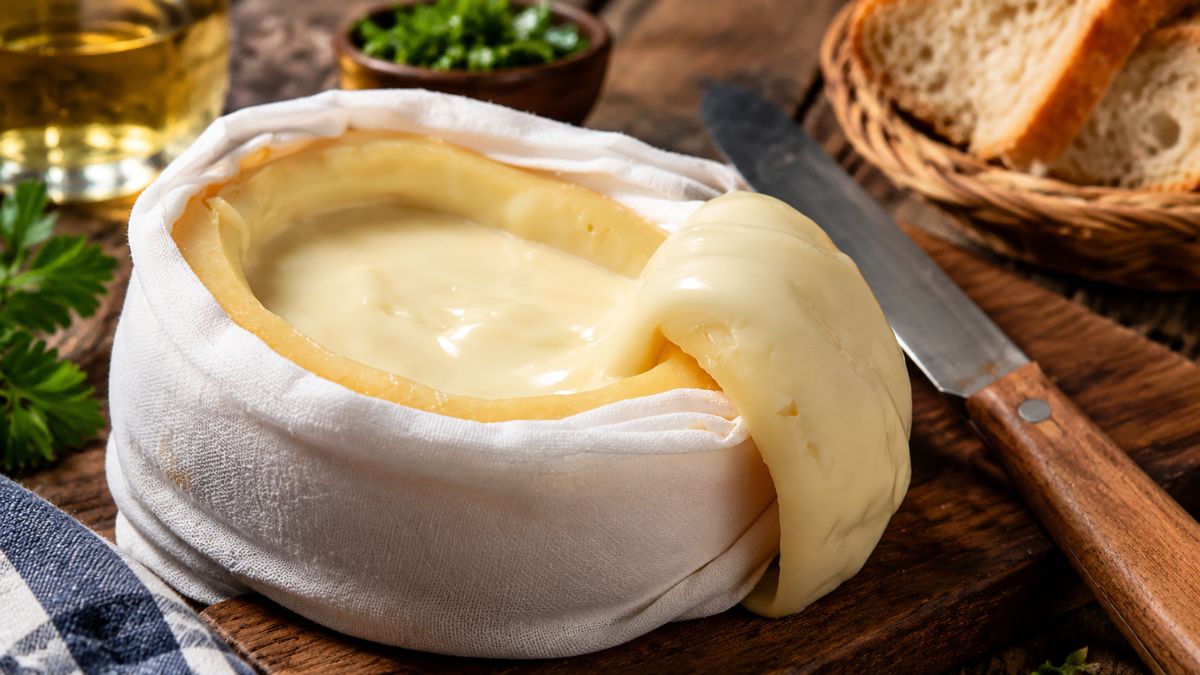 Um Queijo Serra da Estrela artesanal aberto sobre uma tábua de madeira, envolto num pano branco de linho, revelando a sua pasta semimole amanteigada e cremosa a escorrer para o lado. Uma faca está ao lado e um cesto com pão rústico está ao fundo.