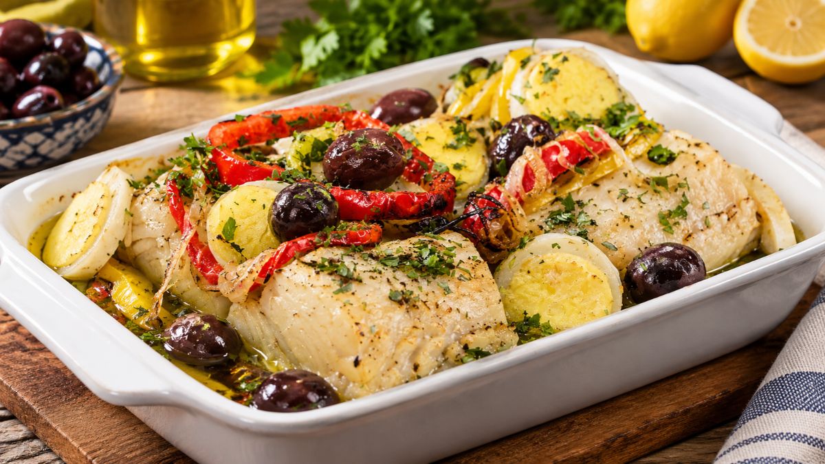 Travessa de vidro com Bacalhau à Gomes de Sá, postas altas sobre batatas e ovos às rodelas, cebola, pimentos vermelhos e azeitonas pretas, polvilhado com salsa fresca.